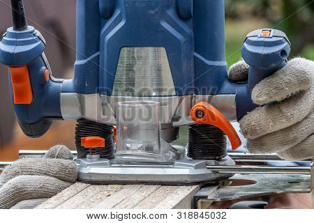 Man In Working Gloves Cuts A Plywood Plate Edge Using A Blue Electric Milling Cutter Tool With Orang