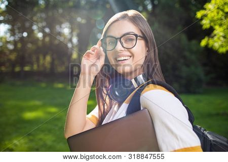 Portrait Of Cute Student Girl With Laptop And Daypack Adjusting Glasses In Park