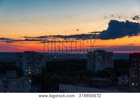 Buildings In The Town Outskirts At Dusk. Low Rise Houses Standing Among Trees Crowns. Lights In Wind