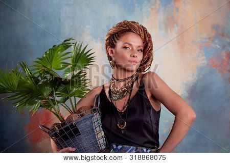 Southern Flavor. A Young Tanned Woman With Afro Braids Smiles Holding A Big Pot With A Tropical Plan