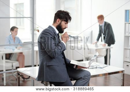 Side view portrait of handsome bearded businessman holding laptop while keaning on desk in modern office, copy space