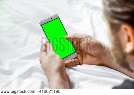 Young Bearded Man Using Green Chroma Key Screen Phone, Relaxing In A Bed. Guy Uses Green Mock-up Scr