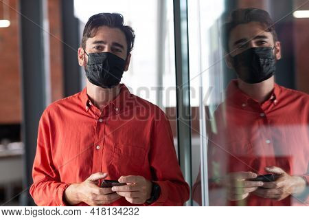 Portrait of caucasian businessman wearing face mask standing in corridor using smartphone. independent creative business during covid 19 coronavirus pandemic.