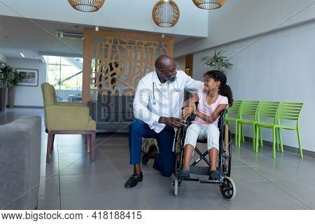 African american male doctor kneeling to talk with mixed race girl in wheelchair in hospital foyer. medicine, health and healthcare services.