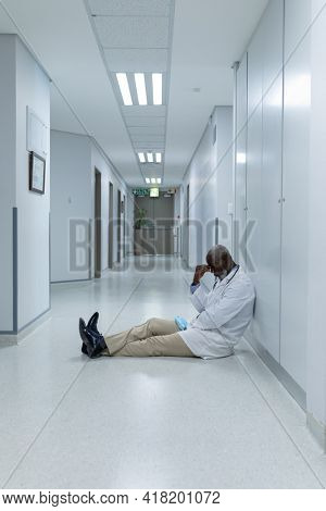 African american male doctor sitting on floor in hospital corridor and worrying. medicine, health and healthcare services.