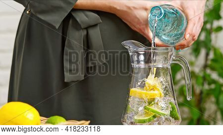 Drink Water. Woman's Hand Pouring Fresh Pure Water With Pieces Of Lemon From Pitcher Into Glass In T