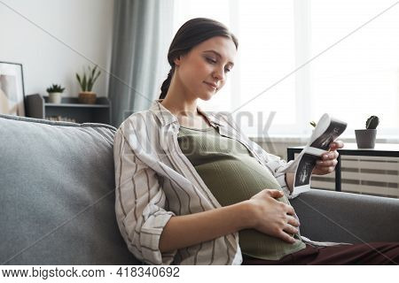 Pregnant Woman Sitting On Sofa And Looking At Ultrasound Image Of Her Baby In The Room