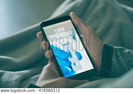 a young man in pajamas, lying on his sofa wrapped in a blanket, checking the result of his covid test in a simulated app in his smartphone, that reads covid test result, and shows a negative result