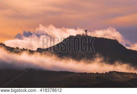 Rocca di Radicofani main tower fortification covered with morning mist. Sunrise light covering the curly clouds running over meadows and fields making light-shadows playing on Tuscany hills landscape.