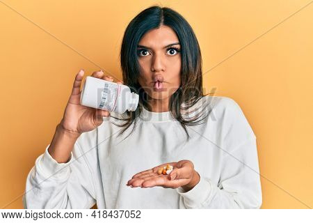 Young latin transsexual transgender woman holding pills making fish face with mouth and squinting eyes, crazy and comical. 