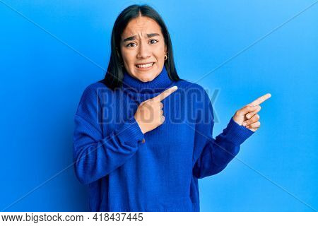 Young asian woman wearing casual winter sweater pointing aside worried and nervous with both hands, concerned and surprised expression 