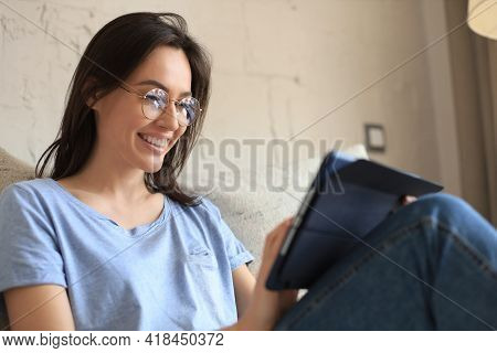 Smiling Young Woman Sitting On Sofa With Digital Tablet And Chating With Friends.