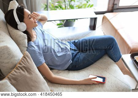 Beautiful Brunette Woman In Earphones Sitting On Sofa Listening To Music With Closed Eyes.