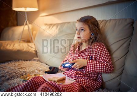 Happy Little Girl In Pajams Sitting On Sofa, Eating Pizza And Pl