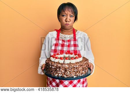 Middle age african american woman wearing baker apron holding homemade cake puffing cheeks with funny face. mouth inflated with air, catching air. 