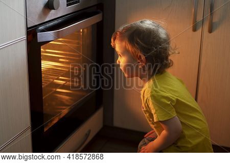A Child Is Sitting Near The Oven In The Kitchen And Waiting. Curious Boy Is Watching Through The Gla