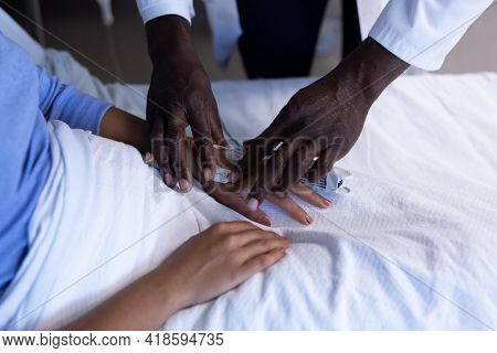 Midsection of african american male doctor putting oximeter on finger of patient in hospital bed. medicine, health and healthcare services.