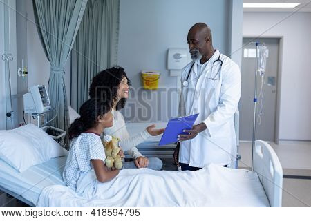 African american male doctor talking with mixed race mother and her sick daughter, in hospital bed. medicine, health and healthcare services.
