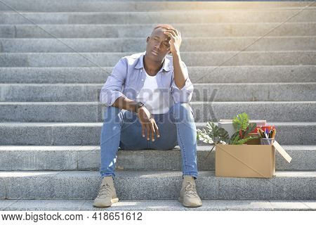 Crisis And Unemployment. Stressed Black Guy With Personal Stuff Sitting On Stairs After Losing His J