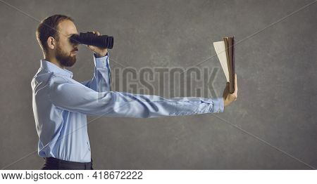 Side View Of Young Guy With Binoculars Reading Book He Is Holding In Long Extended Arm