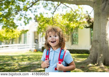 Pupil Of Primary School With Book In Hand Near School Park. Schoolboy With Backpack Near Building Ou