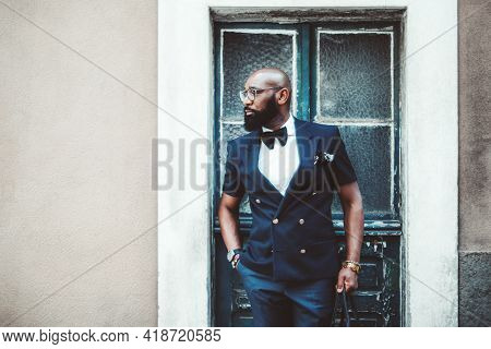 A Handsome Dapper Bald Black Guy With A Beard, In An Elegant Dark-blue Suit With A Bow-tie And Glass