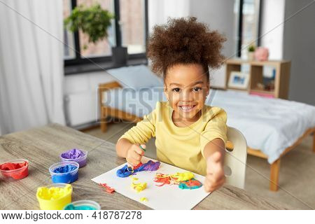 childhood, creativity and hobby concept - little african american girl playing with modeling clay and showing thumbs up at home