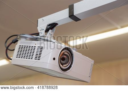 A White Video Projector Hangs From The Ceiling With A Long Arm. The Fluorescent Lamps Are On At The 
