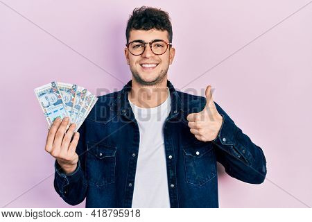 Young hispanic man holding peruvian sol banknotes smiling happy and positive, thumb up doing excellent and approval sign 