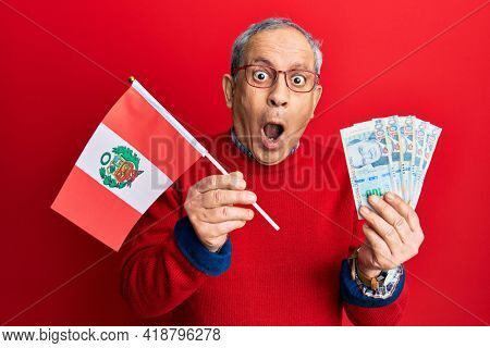 Handsome senior man with grey hair holding peru flag and peruvian sol banknotes afraid and shocked with surprise and amazed expression, fear and excited face. 
