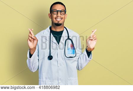 Young african american man wearing doctor uniform and stethoscope gesturing finger crossed smiling with hope and eyes closed. luck and superstitious concept. 