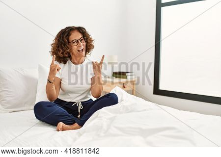 Middle age hispanic woman sitting on the bed at home shouting with crazy expression doing rock symbol with hands up. music star. heavy concept. 