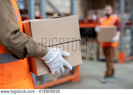 Close-up of unrecognizable mover in gloves and orange vest carrying two boxes while working at warehouse