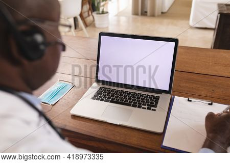 African american senior male doctor writing on clipboard with laptop with copy space on table. distant communication and telemedicine consultation concept.