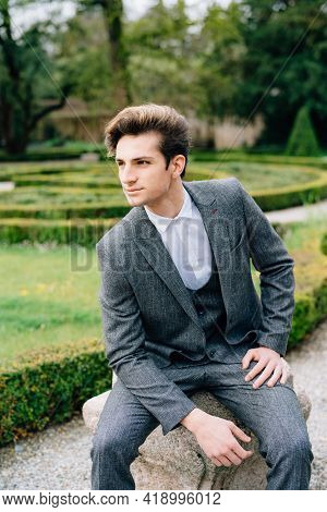 Serious Groom Sits On A Stone In The Park Against The Backdrop Of Trimmed Bushes. Lake Como