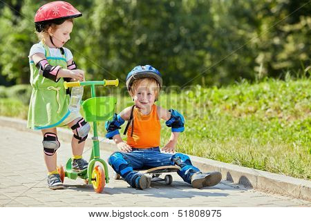 Little boy sits on skateboard with his arms akimbo ,little girl with three-wheeled scooter stands next to him