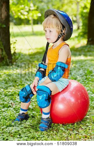 Little boy in protective equipment sits on red ball for jumping on lawn in park