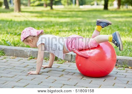 Little girl plays with red ball for jumping on  walkway in park, hands on ground, legs on ball