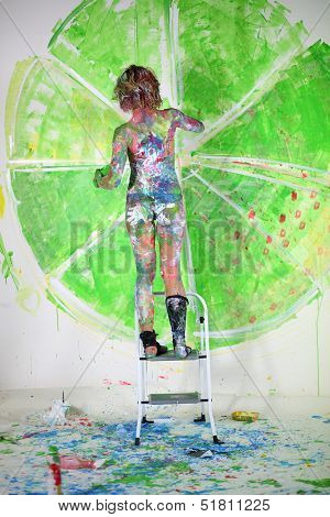 Naked girl covered in paint standing on ladder in studio and draws the wall