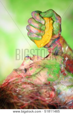 Mans hand squeezes out juice of an orange in mouth of girl covered in paint