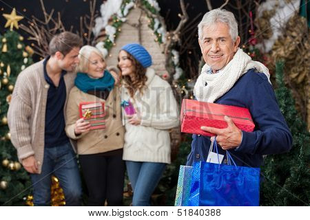 Portrait of happy senior man holding Christmas presents with family standing in background at store