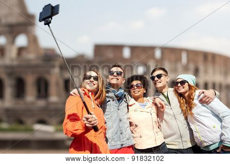tourism, travel, people, leisure and technology concept - group of smiling teenage friends taking selfie with smartphone and monopod over coliseum ruins in rome background