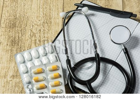 stethoskope on wooden desk with pills pencil and notebook copy space