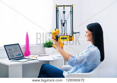 Magic invention. Pleasant beautiful smiling delighted woman sitting at the table and holding model printed on the 3dprinter while expressing gladness