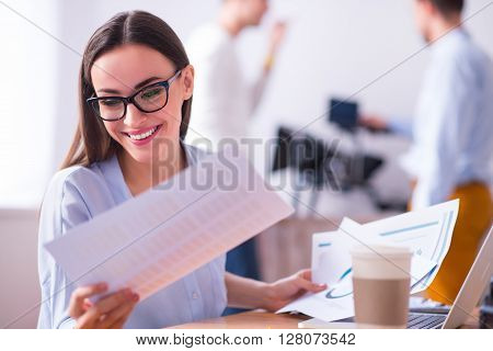 Have a good mood. Cheerful beautiful delighted smiling woman sitting at the table and working with papers while her colleagues standing in the background