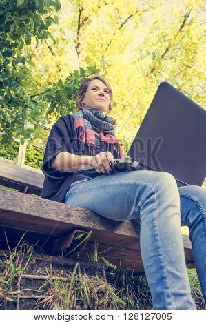 Young woman using laptop at a riverbank. Milleanial working in a park.