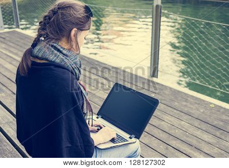 Young woman using laptop at a riverbank. Milleanial working in a park.