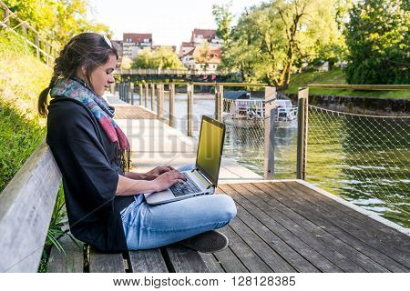 Young woman using laptop at a riverbank. Milleanial working in a park.
