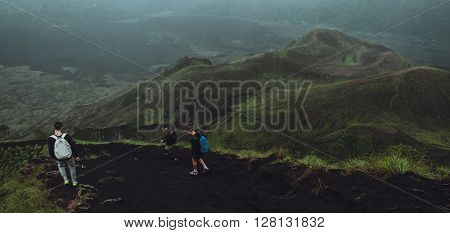 3 Men walk along the hill with backpacks and with white clouds and peak of volcano on background