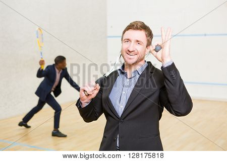 Sporty businessman in business suit smiling for camera and holding ball for squah on court. His business partner playing squash on background.
